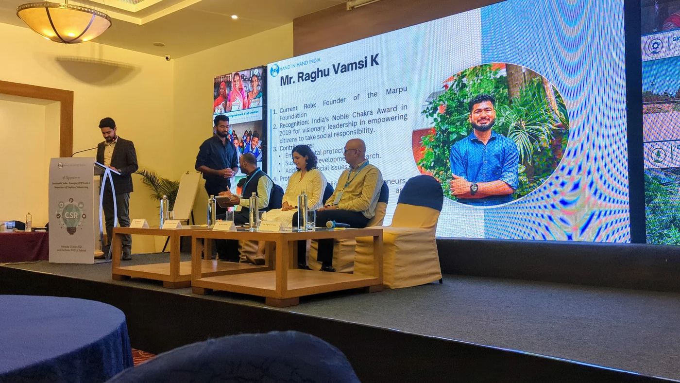 Kadiri Raghu Vamsi speaking at a Hand In Hand India CSR symposium with credentials displayed on stage